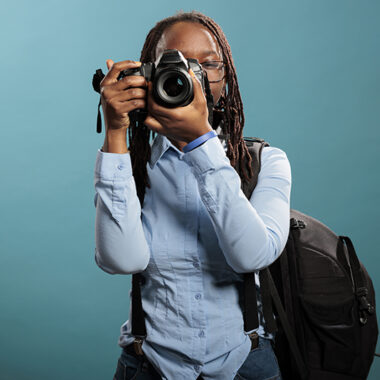 Portrait of professional photographer with photography modern device taking pictures while standing on blue background. Young adult woman with DSLR camera shooting photos. Studio shot